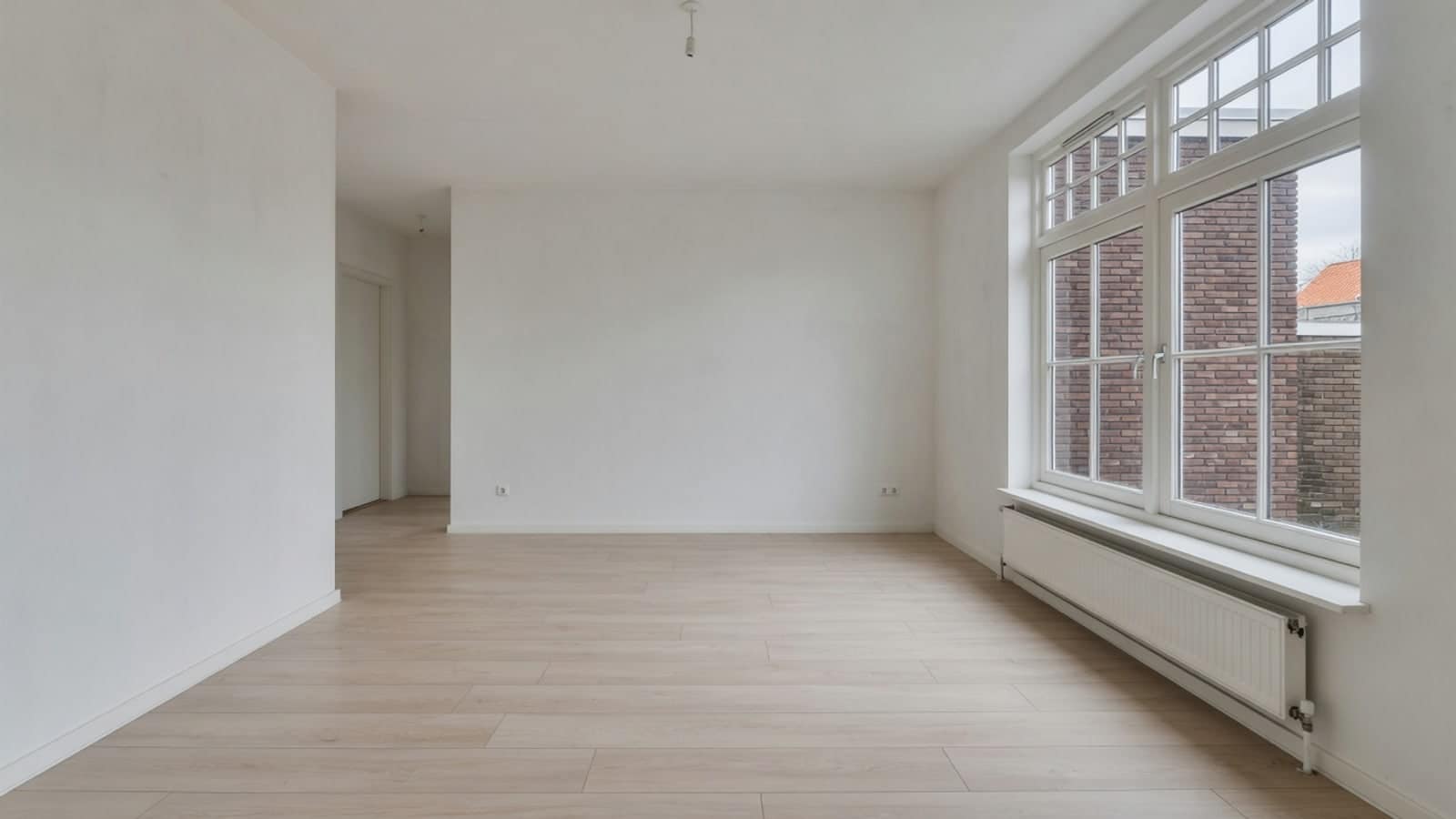Ruime lege woonkamer met een groot vierkant raam en brede lichte vloerdelen